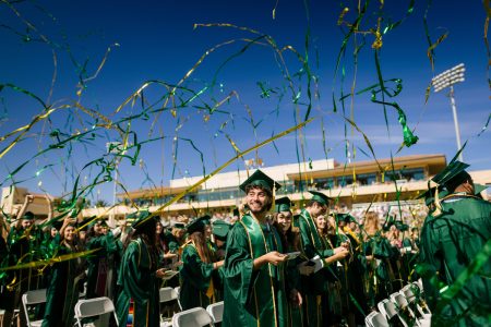 Students celebrate in green caps and gowns at Cal Poly commencement while green and gold streamers fly overhead