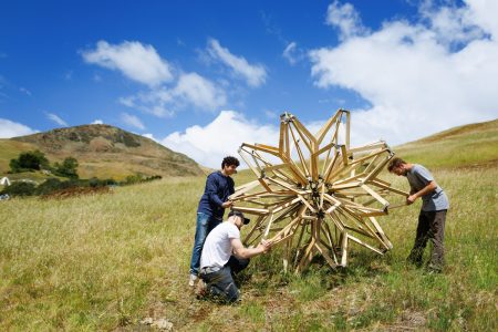 Three students work on a wooden Hoberman's Sphere on a grassy hillside beneath blue sky and clouds
