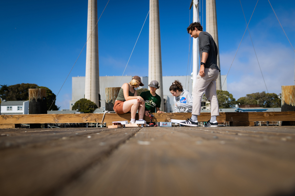 Four students examine hardware on a wooden pier near the smoke stacks of an old power plant in Morro Bay