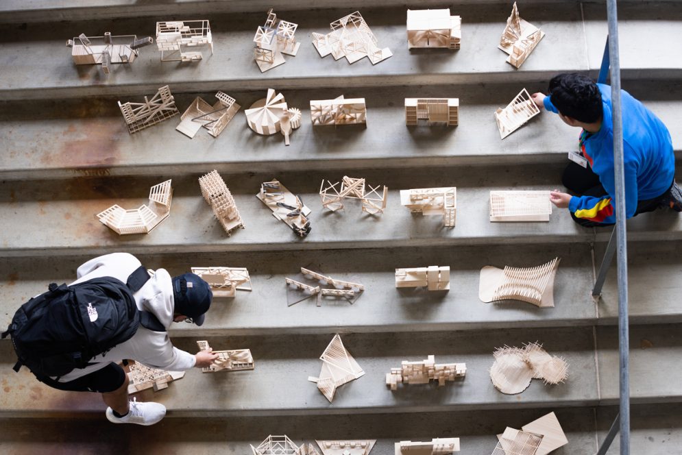 Two students adjust scale models of architectural concepts made of balsa wood on a concrete staircase