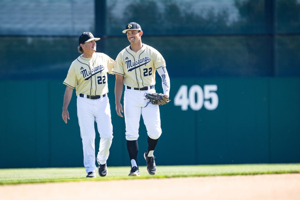 Two people wearing Cal Poly baseball uniforms walk across a baseball field