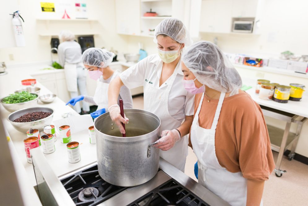 Students wearing hair nets and masks stir a large metal pot over a stove
