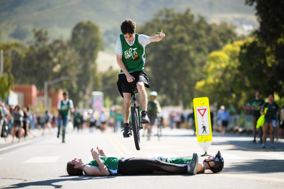 A student riding a unicycle jumps over two people lying on the ground during a parade
