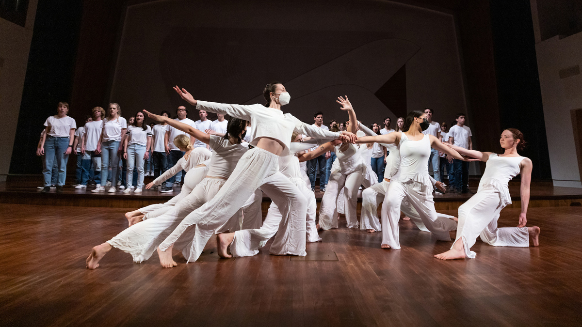 Students wearing white dance in front of a choir in white shirts and jeans on a wooden stage