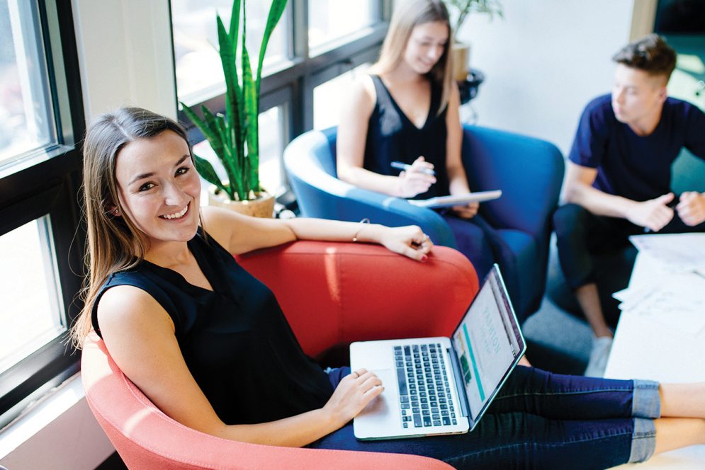 A young woman sits in a chair working on a laptop in front of a bank of sunlight-filled windows, while two other students talk in the background.