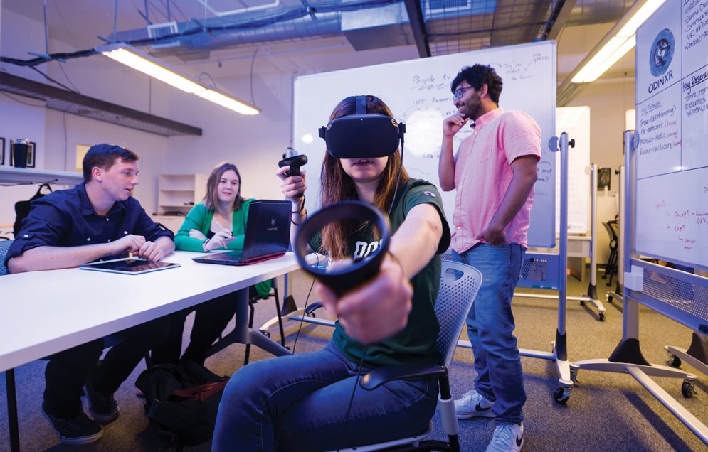 A young woman tests out a VR headset and set of controllers while three other students talk in the background in their workspace.