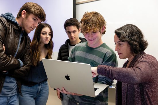Four students and a professor gather around a laptop.