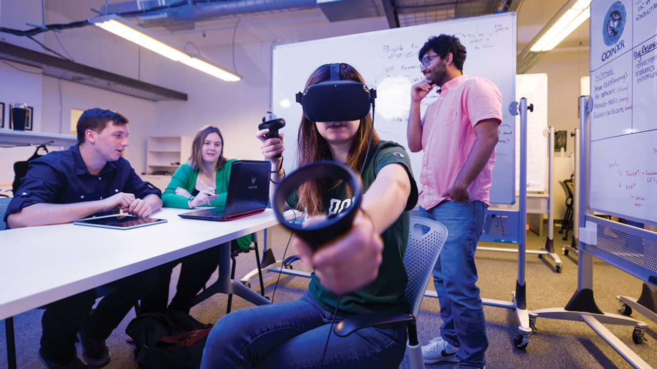 A young women tests out a VR headset and controller while her teammates talk behind her in a workspace surrounded by white boards.