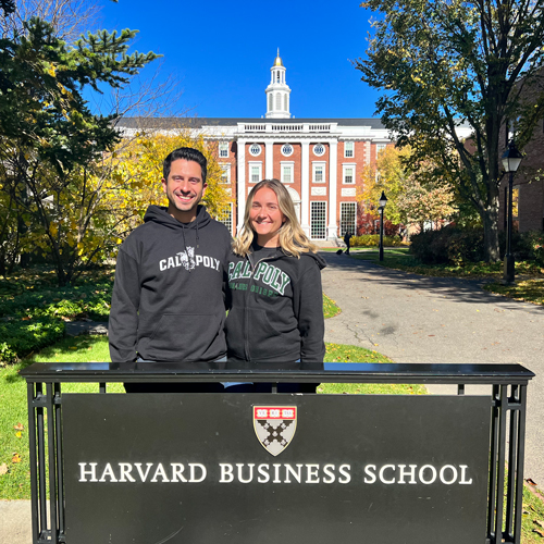 Two people wearing Cal Poly sweatshirts stand in front of a Harvard Business School sign