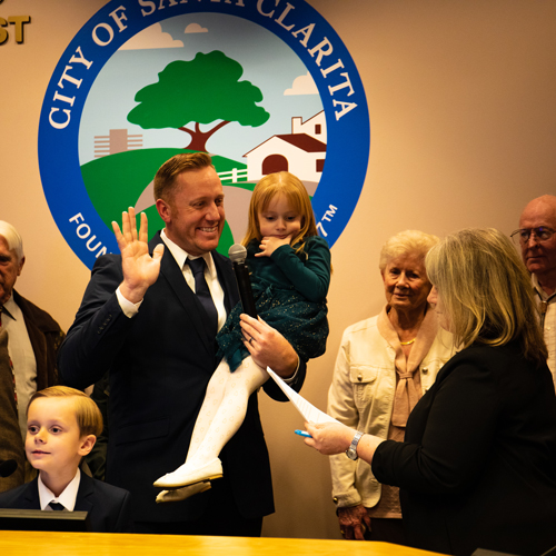 Jason Gibbs holds a young child while raising his hand to be sworn in near the seal of the City of Santa Clarita