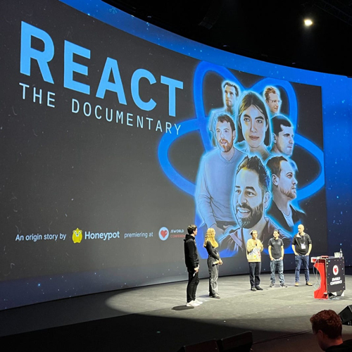 Five people stand on a stage in front of a screen with the words 'React: The Documentary'