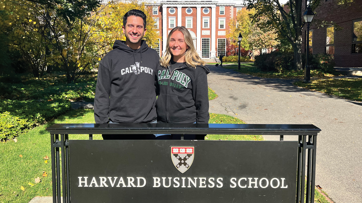 Two people in Cal Poly sweatshirts stand in front of a Harvard Business School sign