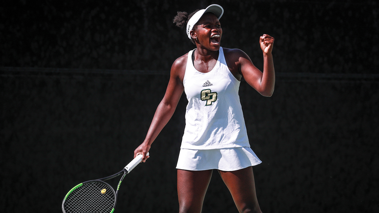 A tennis player in a white visor and dress holds a tennis racket while celebrating.