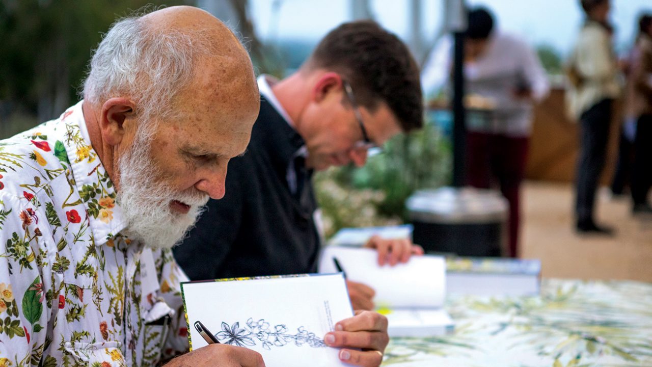 Two people sit at a table and sign open books with images of flowers on the pages