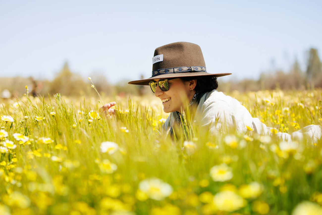 A student in a wide brimmed hat and sunglasses lays in a field of yellow wildflowers, inspecting one up close.