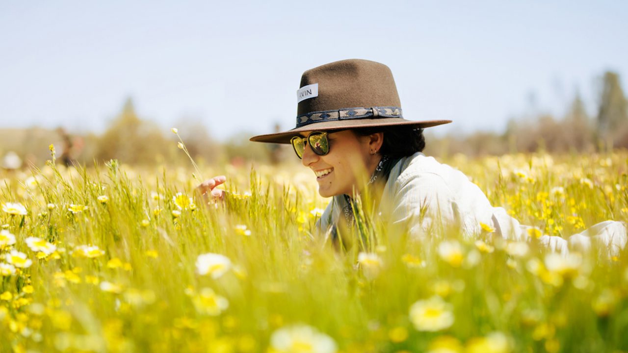 A student in a wide brimmed hat and sunglasses lays in a field of yellow wildflowers, inspecting one up close.