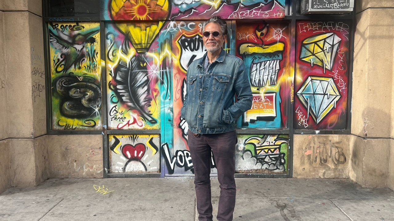A man in sunglasses stands before a wall with graffiti