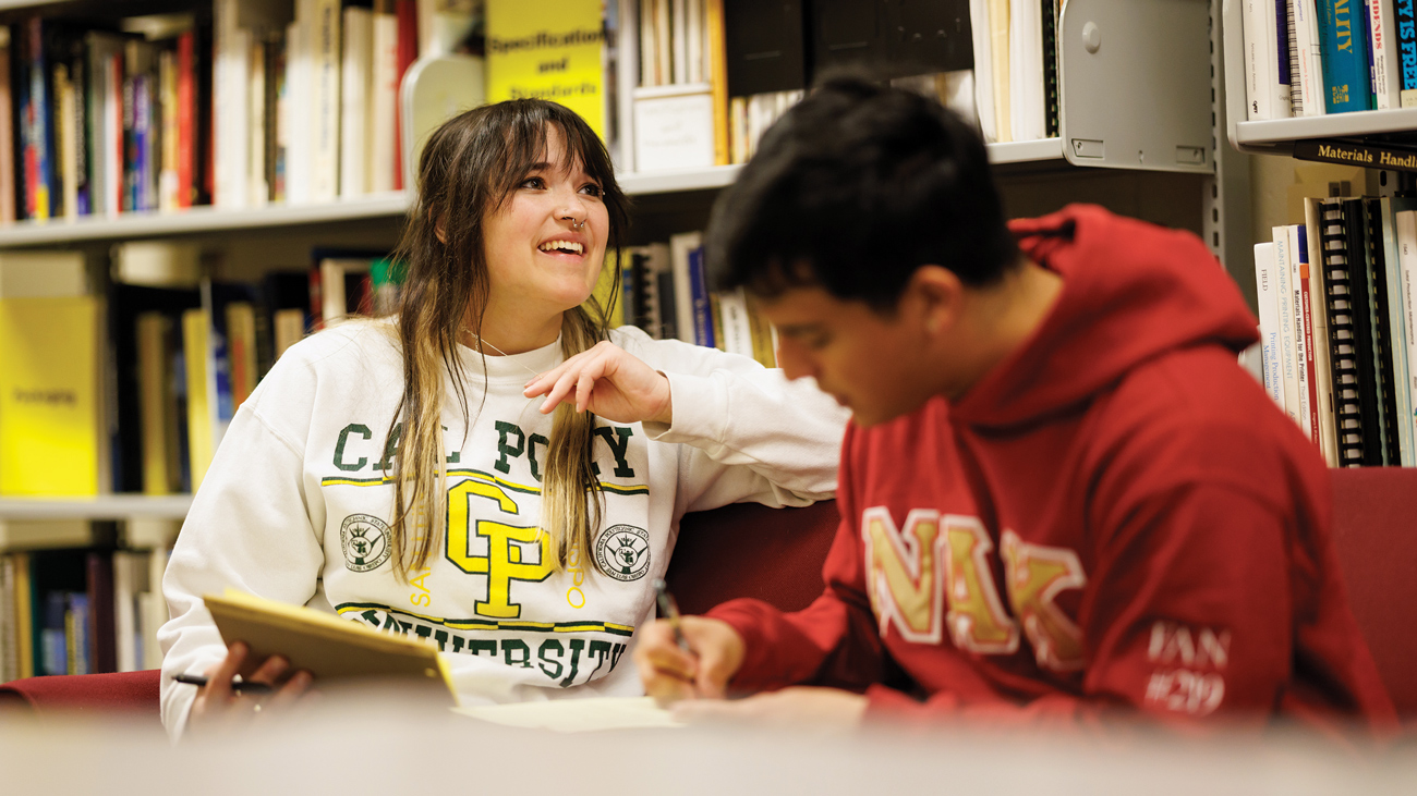 Two students hold notepads and collaborate near shelves of books before a debate exhibition at Cal Poly