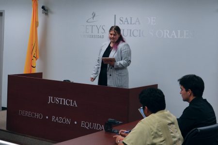 A student wearing a blazer reads from a notepad during a Spanish language debate near a podium that reads 'Justicia, Derecho, Razón, Equidad'