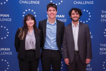 Three students smile in front of a blue backdrop that reads Schuman Challenge