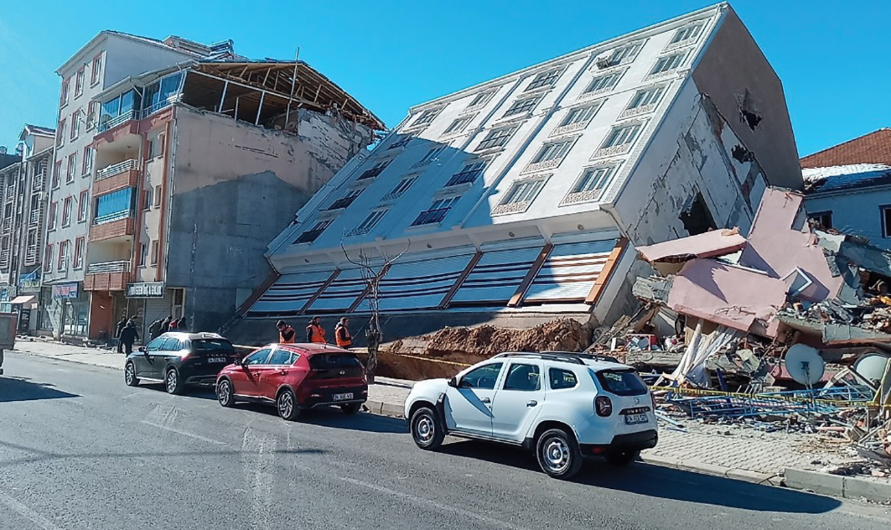 Workers inspect the damage as a five-story building lays at an angle away from a Turkish street, surrounded by rubble.