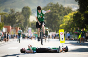 A young man on a unicycle wearing a green jersey jumps over two other students lying on the sidewalk