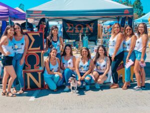 A group of young Latina women pose in front of a booth with Sigma Omega Nu banners