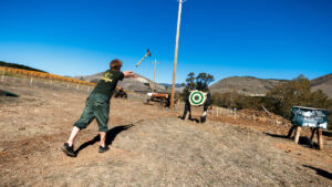 A student in a Cal Poly Logging Team t-shirt throws a double-bit axe at a distant wood target.