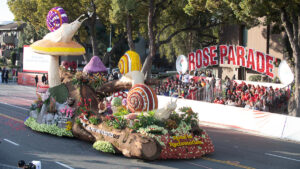 A float featuring snails, flowers and a giant mushroom rolls down a street in front of a sign reading "Rose Parade"