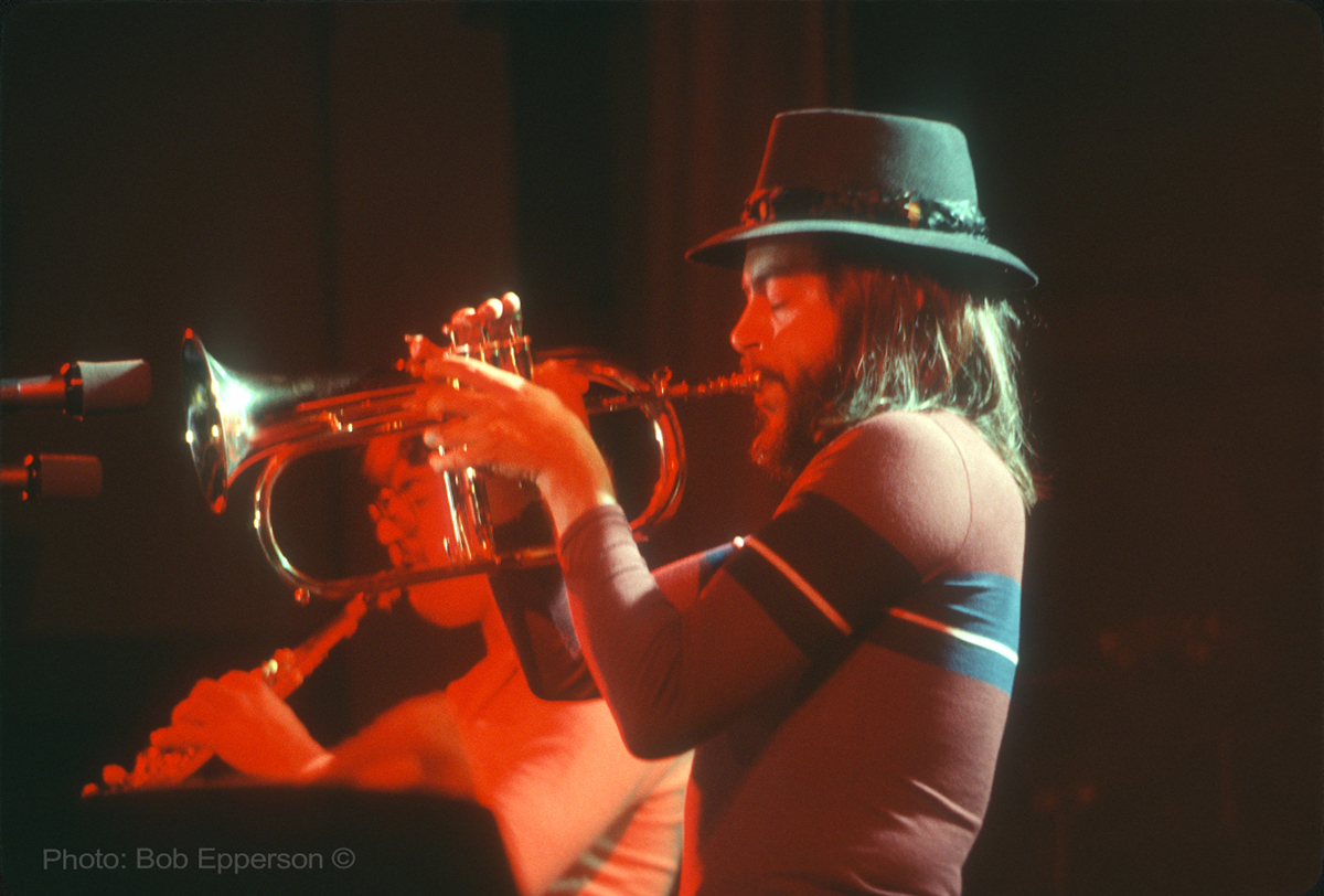 A bearded man in a hat plays a flugelhorn into a microphone.