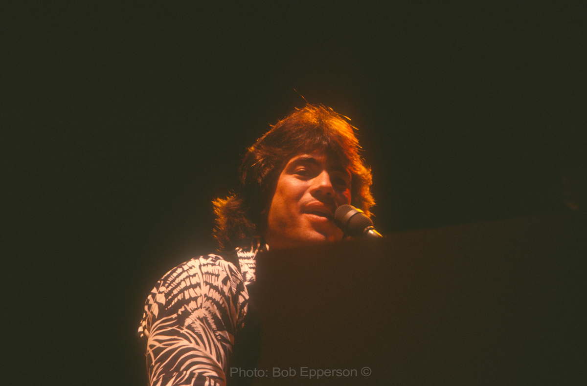 A man with shaggy hair sings into a microphone while playing a keyboard.