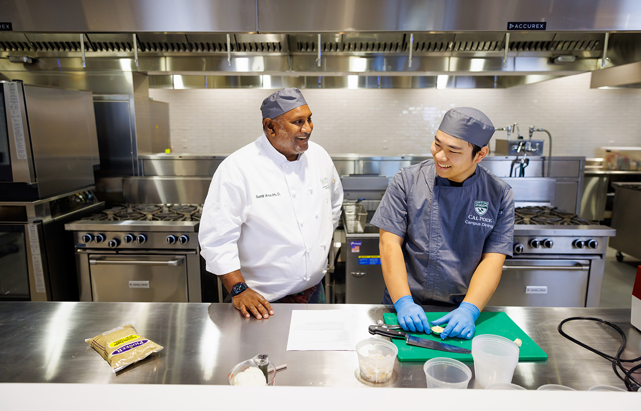 A professor and student in chef coats and caps talk while standing at a kitchen sork station.