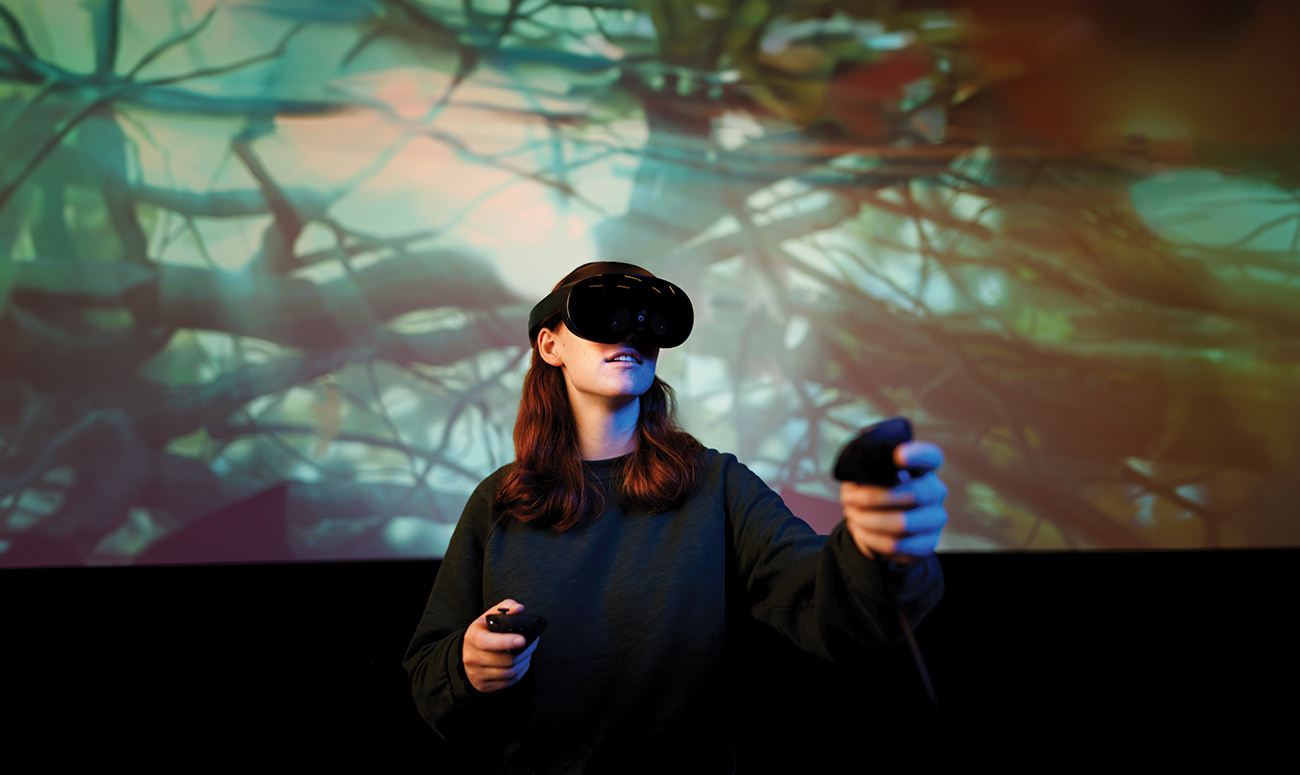 A young women uses VR controllers while wearing a VR headset in front of a screen showing a forest scene.