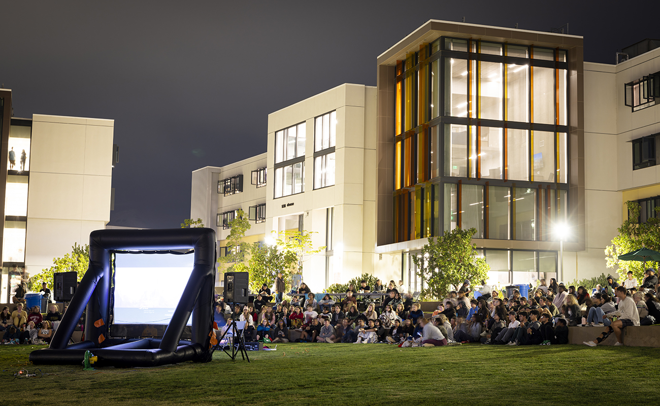 In front of a lit residence hall building after dark, a group of students sits on a lawn watching a movie projected onto a large portable screen.