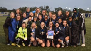 A group of young women in athletic gear pose for a photo on a field, holding a title plaque.