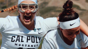 Two young women in Cal Poly lacrosse jerseys and athletic headbands, one wearing an eye protector, laugh and smile as they hug each other on a field