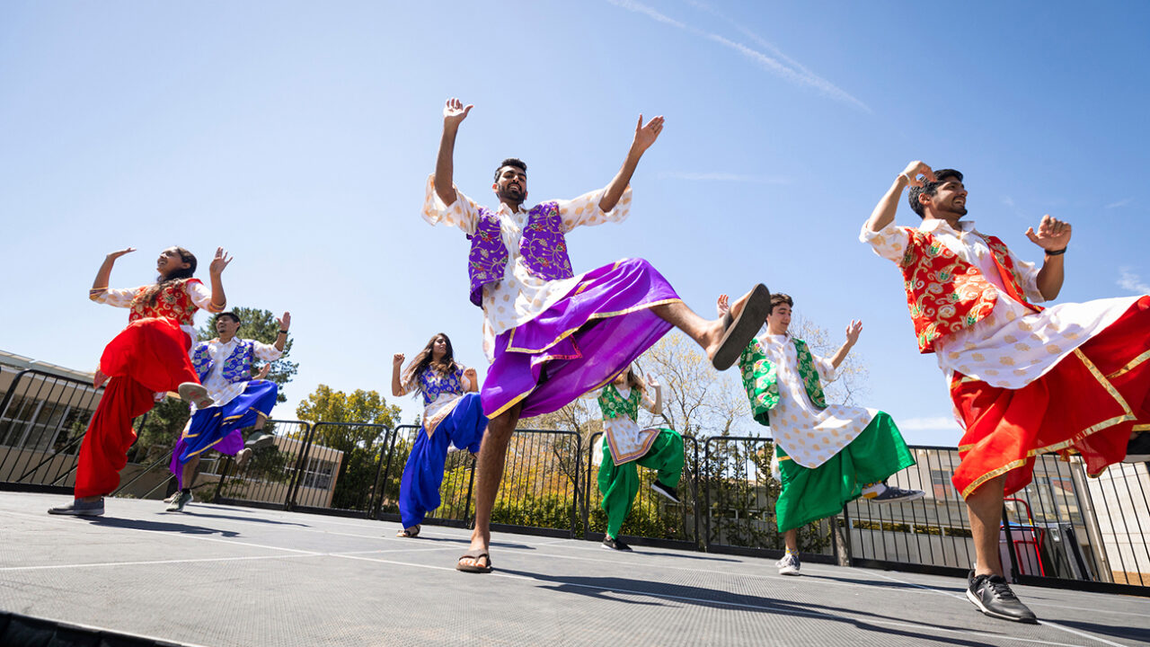 A group of students dressed in traditional Indian costumes perform a dance on an outdoor stage.