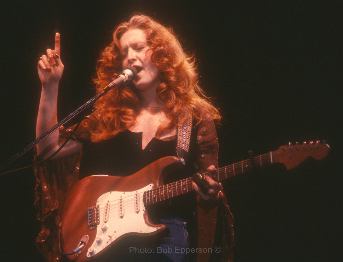 Singer Bonnie Raitt sings into a microphone while holding a red guitar