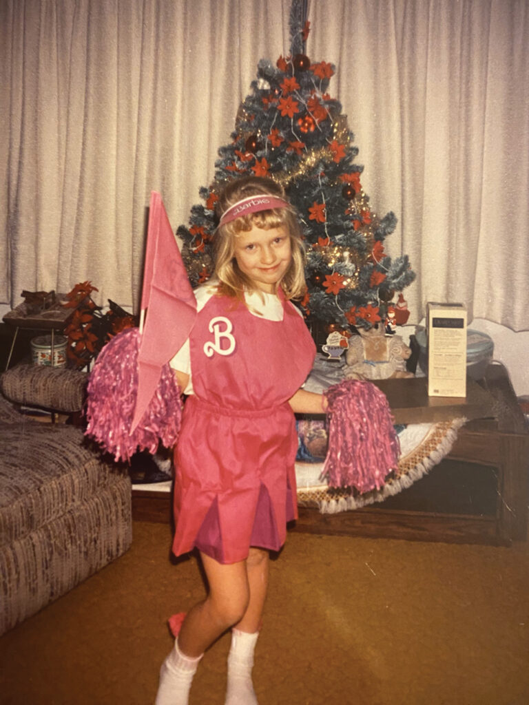 A young girl in a vintage photo wears a pink dress with the Barbie "B" logo and holds Barbie accessories in front of a Christmas tree
