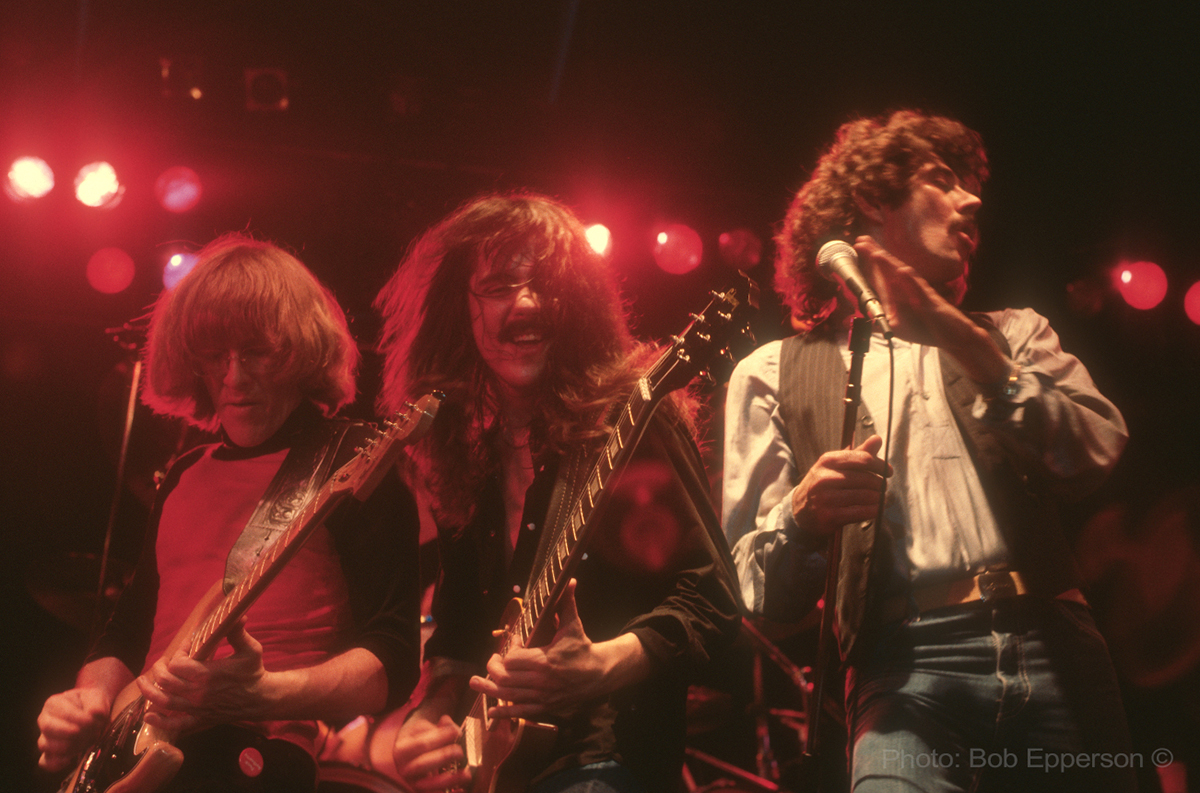 Three backlit men lean toward each other on stage, two playing guitar and one gripping a microphone, in a vintage photo.
