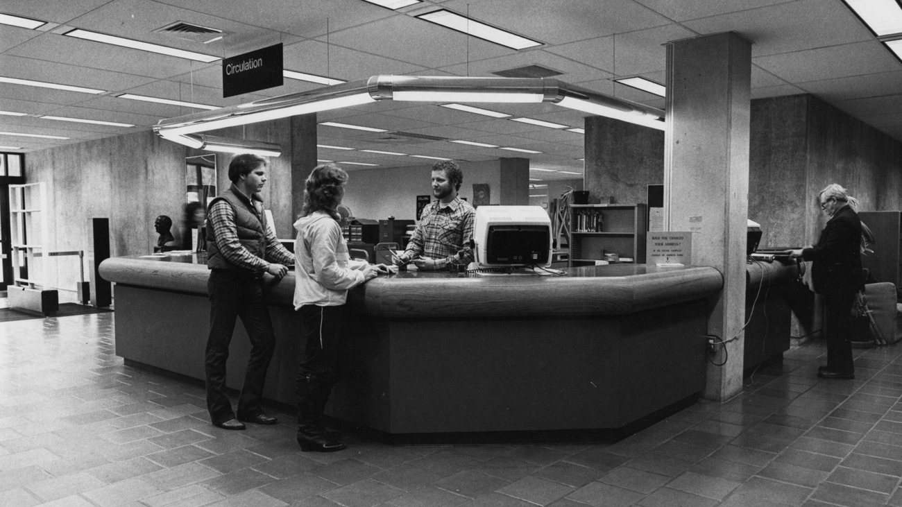 A black and white image of student and staff congregating around Kennedy Library's reception desk