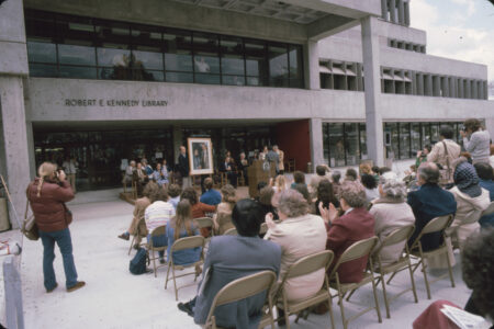 A seated crowd and photographers look toward a podium and speakers in Kennedy Library's front entrance at its dedication ceremony.