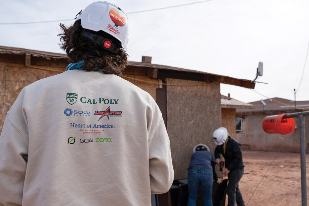 A student wearing a sweatshirt with Cal Poly and Heart of America logos looks at his teammates installing solar panels on a home