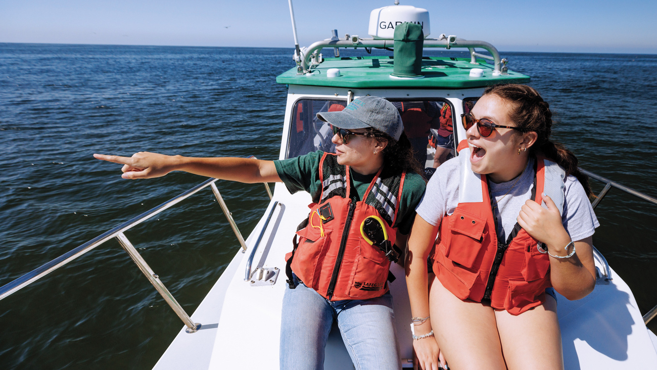 Two students wear life jackets, smile and point while riding on a boat to deploy audio data analysis technology.