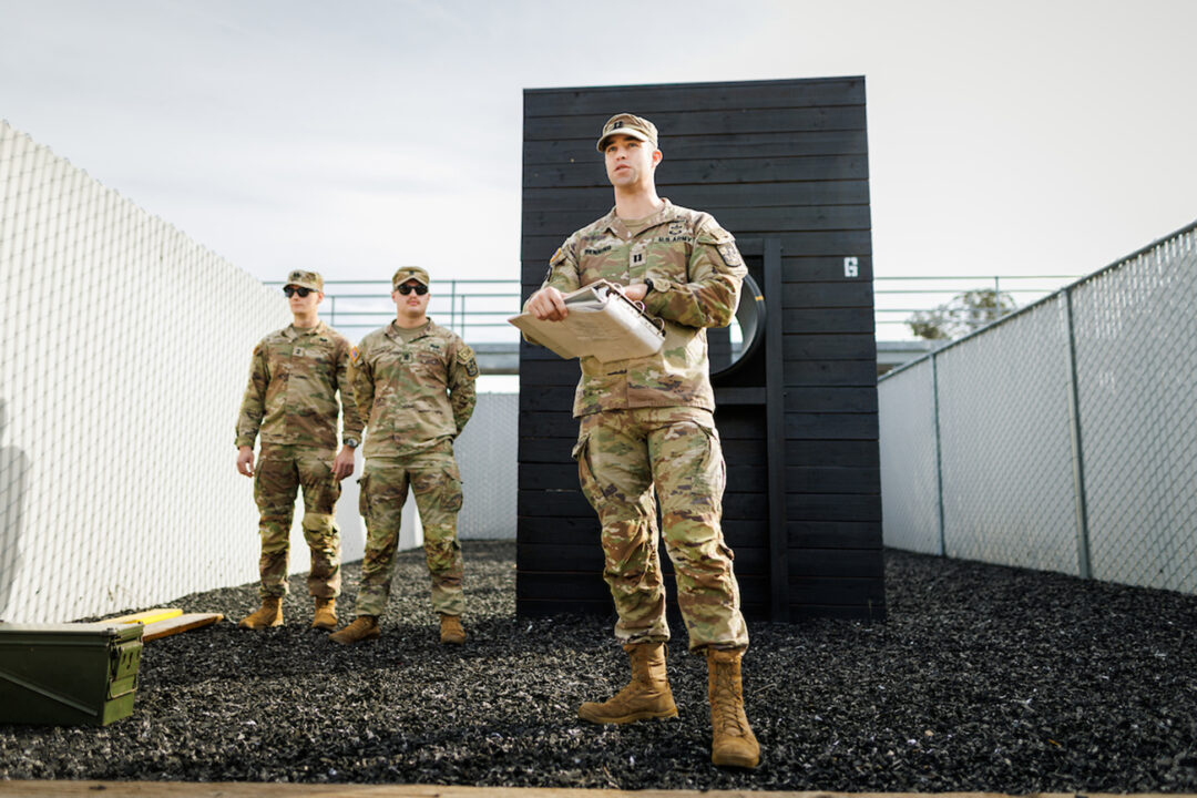 An Army officer in camo fatigues reads from a clipboard, standing in front of an obstacle course and two uniformed cadets.