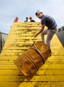 With the support of classmates at the top, a young man uses a rope to haul a barrel up a steep yellow ramp.