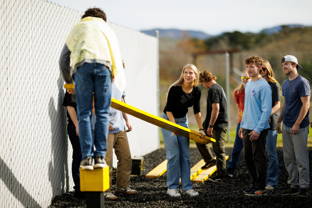 A young man wearing a yellow cape stands on a raised post, handing a long plank to a group of classmates.