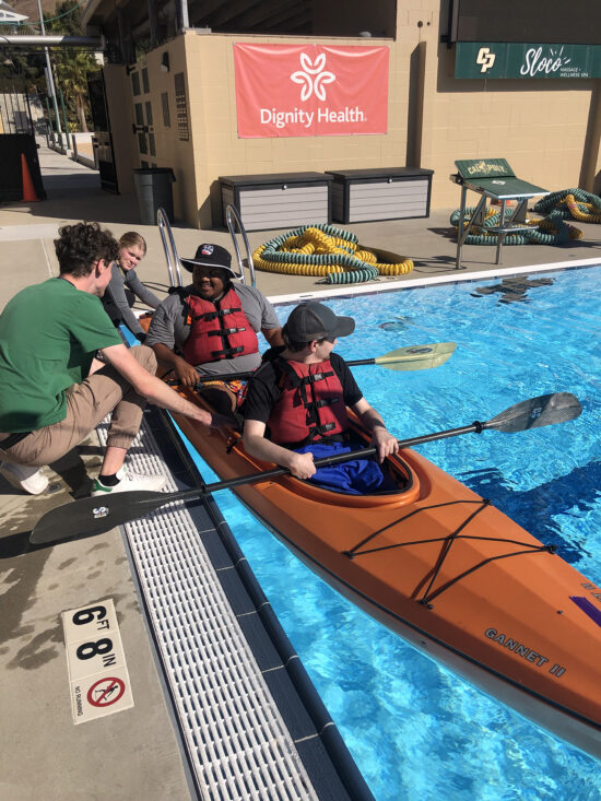 Two students assist kayakers paddling their craft in a swimming pool.