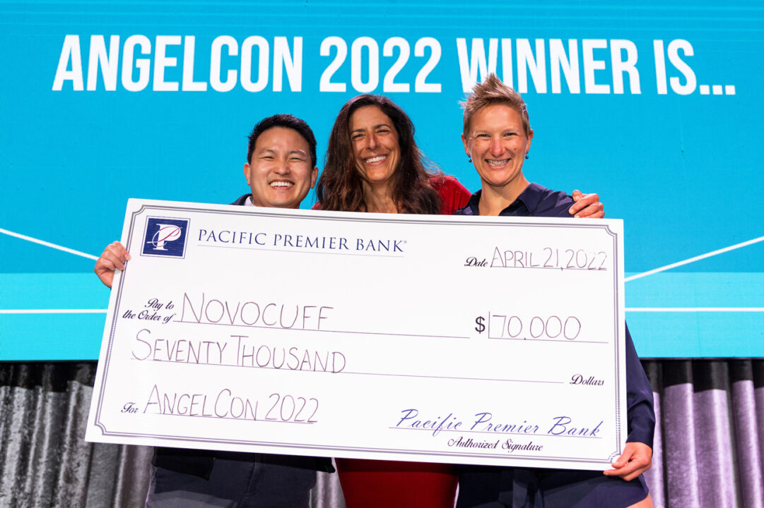 Three people stand behind a $70,000 oversized check on stage near a screen displaying the text Angelcon 2022 winner is