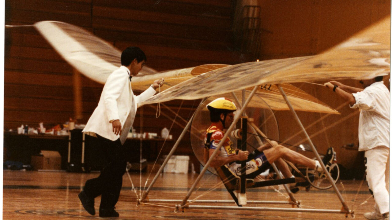 Three people tend to a human-powered helicopter in a gymnasium.